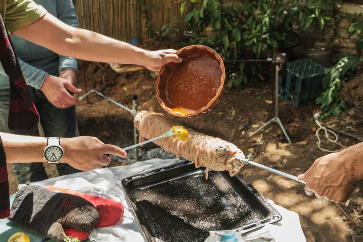 Hands preparing kokoretsi, a traditional Greek dish, by pouring a liquid over it as it cooks on a spit outdoors.